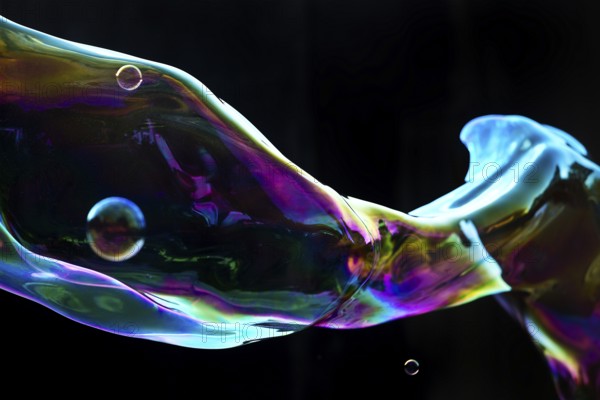 Large, iridescent soap bubble tube in the colours of the rainbow against a black background, Recklinghausen, Germany