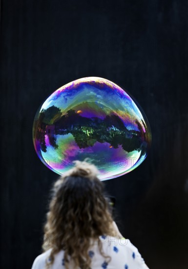 A woman looks at a large iridescent and floating soap bubble in the colours of the rainbow against a black background, Recklinghausen, Germany