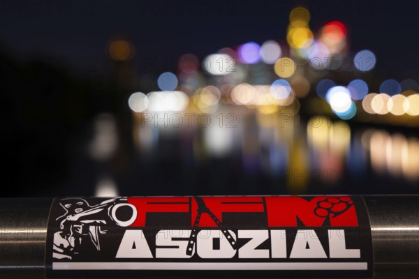 The lights of Frankfurt's banking skyline are blurred in the background. In front of it, the sticker FFM ASOZIAL is stuck on a bridge railing. Frankfurt am Main is repeatedly counted among the cities with the highest crime rate, Osthafen, Frankfurt am Main, Hesse, Germany