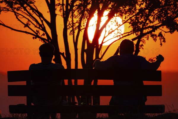 From the summit plateau of the GroÃŸer Feldberg in the Taunus mountains, two people watch from a bench as the sun sets on the distant horizon, Hesse, Germany