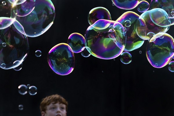 A boy marvels at shimmering and floating soap bubbles against a black background, Recklinghausen, Germany