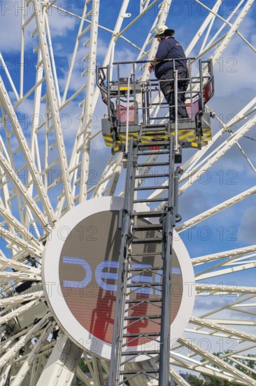 A worker stands on a ladder in front of a large Ferris wheel under a blue sky, fire brigade exercise for gondola rescue Ferris wheel, Calw, Germany