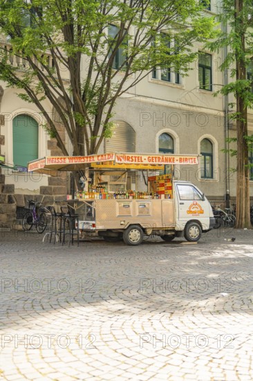 A food truck stands under trees on a paved square on a sunny day, Merano, South Tyrol, Italy