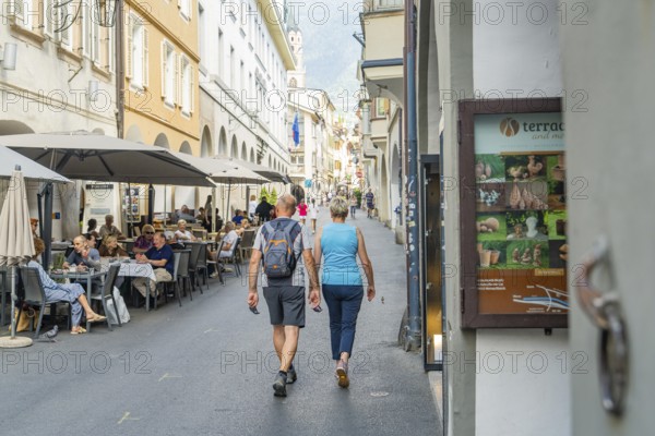 Couple walking through a pedestrian zone with restaurants in the background, mountain view in summer, Merano, South Tyrol, Italy