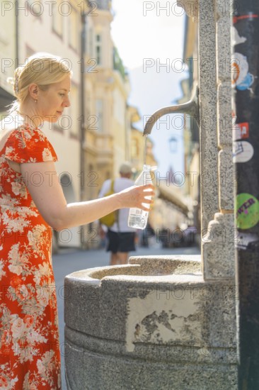 Woman filling a water bottle at an old fountain in the city, Merano, South Tyrol, Italy