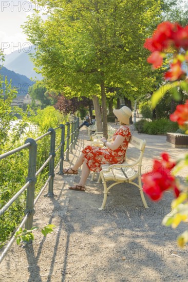 Woman in summer dress sitting on park bench, surrounded by flowers in relaxed atmosphere, Merano, South Tyrol, Italy