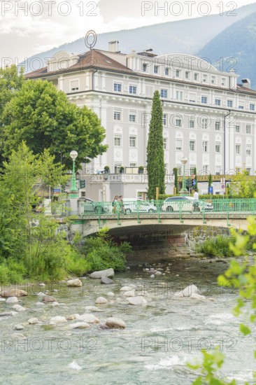 Historic riverside hotel with bridge and lush vegetation against a mountain backdrop, Merano, South Tyrol, Italy