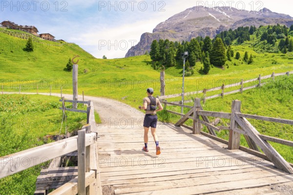 A runner crosses a wooden bridge in a green, mountainous landscape under a blue sky, Zallinger Alm, Dolomites, Italy
