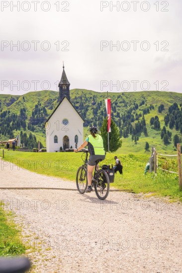 A cyclist rides along a gravel path, in the background a village church and green hills, Zallinger Alm, Dolomites, Italy
