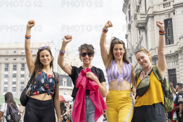 Four women are raising their arms, wearing rainbow bracelets and smiling, celebrating lgbtq rights at a gay pride parade, showing unity and demanding equality