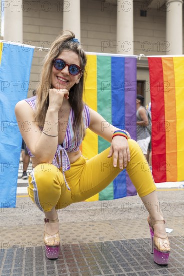 Young woman crouching and smiling brightly, wearing stylish sunglasses and vibrant, colorful clothes, celebrating gay pride with rainbow flags waving in the lively background