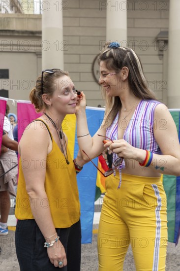 During a vibrant gay pride event, a woman applying make-up to her friend's face highlights the joyous atmosphere of celebration, inclusivity, and community spirit
