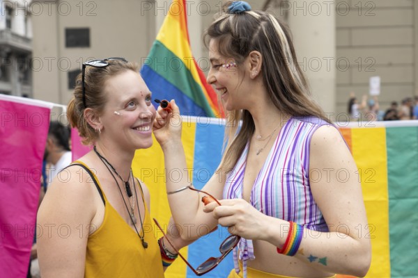 Two women are smiling and putting rainbow makeup on their faces at a gay pride parade, celebrating diversity and lgbtq rights, surrounded by colorful flags and a joyful atmosphere