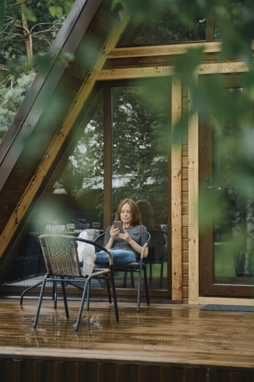Serene moment after the rain. Pretty woman is reading something on smartphone. She is seated comfortably outside a modern wooden cabin with large glass windows, surrounded by lush greenery