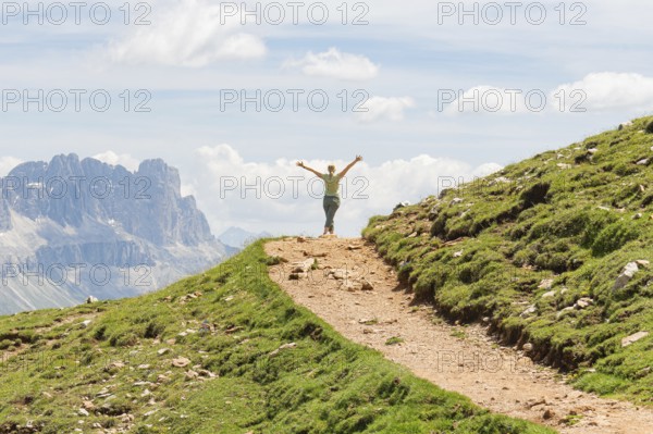 Person with outstretched arms on a path with a wide mountain view, Schlehrn hike, Alpe di Siusi, Dolomites, South Tyrol, Italy