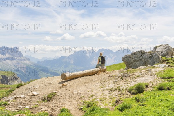 Hiker sitting on a tree trunk with a wide view over the mountains and valleys, Schlehrn hike, Alpe di Siusi, Dolomites, South Tyrol, Italy