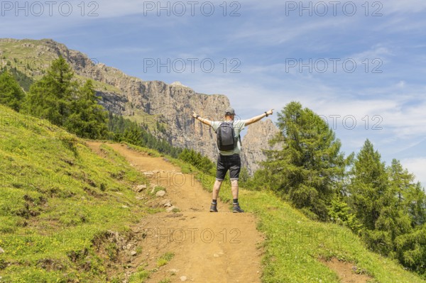 A hiker with outstretched arms on a mountain path, surrounded by green nature, Schlehrn hike, Alpe di Siusi, Dolomites, South Tyrol, Italy