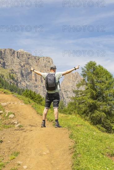 A man stands joyfully with outstretched arms on a sunny mountain path, Schlehrn hike, Alpe di Siusi, Dolomites, South Tyrol, Italy