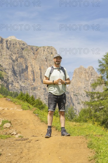 A man with a rucksack stands smiling on a mountain path with a rocky background, Schlehrn hike, Alpe di Siusi, Dolomites, South Tyrol, Italy