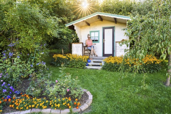 A woman sits relaxed on the small veranda of a garden shed and reads. Blooming summer flowers in the garden. Germany