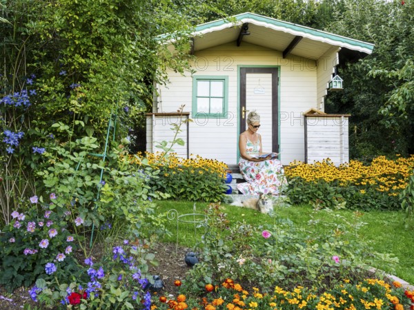 A woman in a dress sits relaxed on the small veranda of a garden shed and reads. Blooming summer flowers in the garden. Germany