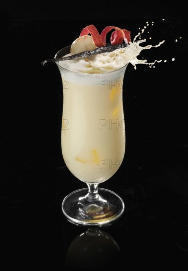 Cocktail in a highball glass on ice cube with lychee and vanilla pod as garnish against a black background, splash of ice cubes thrown in, studio shot
