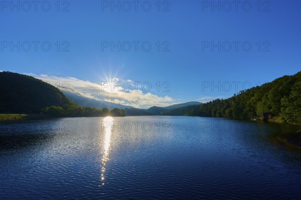 Lake in the morning light with sun, blue sky and surrounding mountains, summer, Rursee, Obersee, Rurberg, Simmerath, Eifel National Park, Eifel, North Rhine-Westphalia, Germany