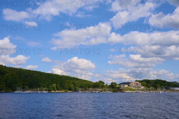 A lake with a building and boats along a wooded shore under a cloudy sky, summer, Rursee, Rurtalsperre, Heimbach, Simmerath, Eifel National Park, North Rhine-Westphalia, Germany