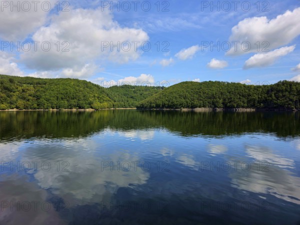 Blue sky with white clouds and their reflection in a calm lake, surrounded by lush forests, summer, Rursee, Rurtalsperre, Simmerath, Eifel National Park, North Rhine-Westphalia, Germany