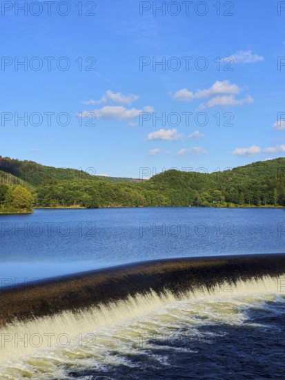 A quiet reservoir with a small waterfall, surrounded by wooded hills under a blue sky, summer, Rursee, Obersee, Rurberg, Simmerath, Eifel National Park, North Rhine-Westphalia, Germany