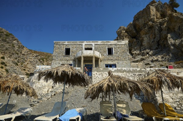 Stone house against a rocky backdrop with beach loungers and thatched roofs in the sunshine, Embros Thermal Baths, Kos, Dodecanese, Greek Islands, Greece