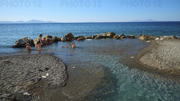 People swimming in the calm blue sea by rocks and sandy shore, Embros Thermal Baths, Kos, Dodecanese, Greek Islands, Greece