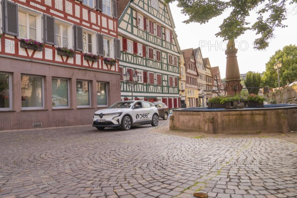 A car next to a fountain in a cobbled old town street with half-timbered facades and green plants, Deer E, car sharing, electric car, Calw, Black Forest