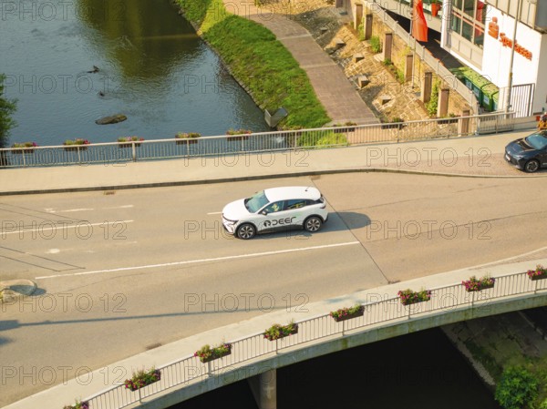 A white car drives on a bridge over a river, surrounded by urban architecture and nature, Deer E, car sharing, electric car, Calw, Black Forest
