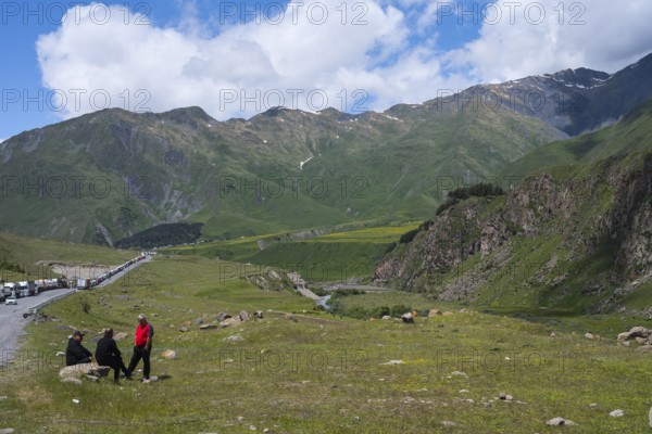 Three people sit in a meadow in front of a mountain range under a cloudy sky, left truck queue on the way to Russia, Kazbegi, Qasbegi, Stepantsminda, Georgian Military Highway, Mtskheta-Mtianeti Province, High Caucasus, Georgia