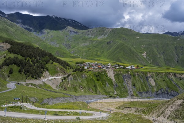 A small village is situated on a hill surrounded by green mountains and cloud-covered sky, views of the Terek River valley and Kanobi, Kazbegi, Kasbegi, Stepantsminda region, Georgian Military Highway, Mtskheta-Mtianeti province, High Caucasus, Georgia