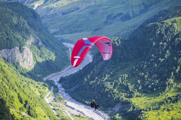 A paraglider floats over a narrow, green valley region with a fast-flowing river, paraglider near Gudauri, White Aragvi River, Mtskheta-Mtianeti region, Georgian Military Highway, High Caucasus, Georgia
