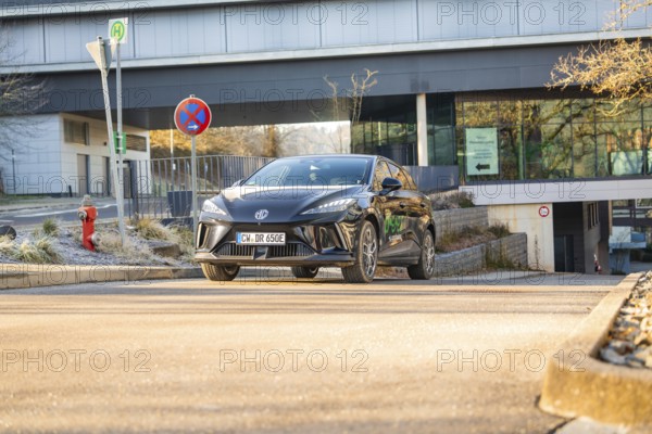 Black car drives in front of a modern building backdrop during the day, road signs show the way, MG4 electric car, deer e-car sharing, Calw, Germany