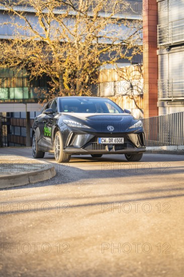 Black car drives on a curved road past an architecturally designed building, MG4 electric car, Deer e-Carsharing, Calw, Germany