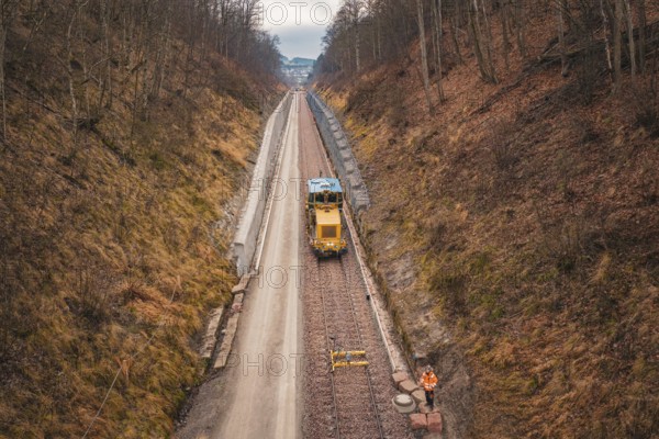 Yellow train and workers on a long railway line through autumnal forest, using a tamping machine at the Hermann Hessebahn construction site, Calw, Germany