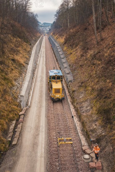 Yellow train on rails with workers in street surrounded by bare forest, using tamping machine at Hermann Hessebahn construction site, Calw, Germany
