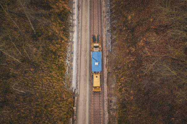 Aerial view of a yellow train traveling on rails surrounded by bare trees, using a tamping machine on the Hermann Hessebahn construction site, Calw, Germany