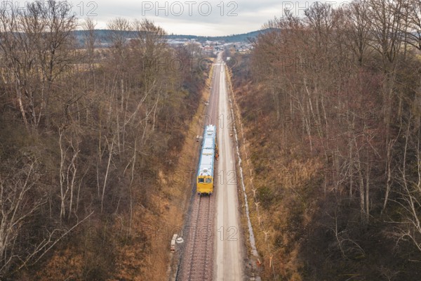 Long view of a rail line with a yellow train through a wintery forest, using a tamping machine at the Hermann Hessebahn construction site, Calw, Germany