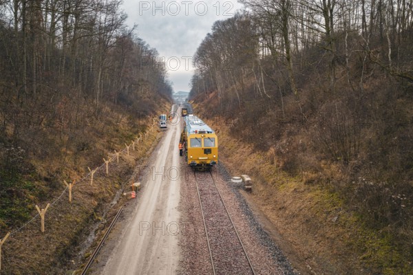 Yellow train on a lonely railway line through bare forest under cloudy sky, using a tamping machine at the Hermann Hessebahn construction site, Calw, Germany