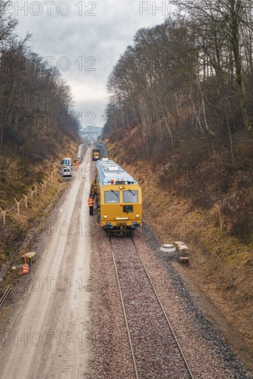 Train with construction workers on a rail line surrounded by winter forest, using a tamping machine at the Hermann Hessebahn construction site, Calw, Germany