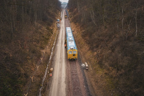 View of a railway line with a yellow train in a lightly wooded area, tamping machine used at the Hermann Hessebahn construction site, Calw, Germany