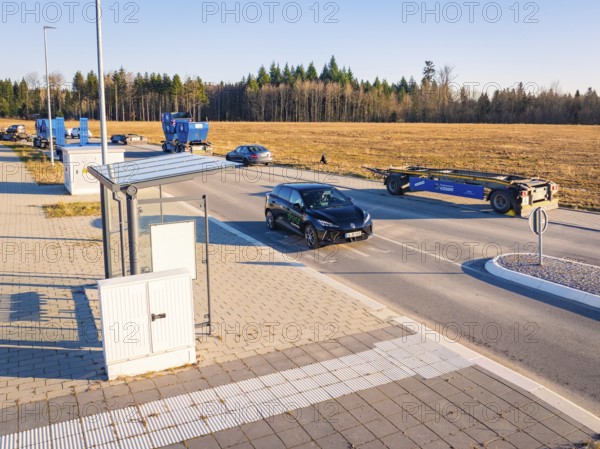 A car turns onto a wide road surrounded by parked vehicles and fields, MG4 electric car, Deer E- Carsharing, Calw, Germany