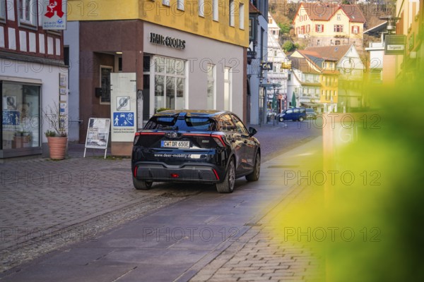 Car driving through a narrow city street surrounded by colorful buildings in the background, MG4 electric car, Deer e-Carsharing, Calw, Germany