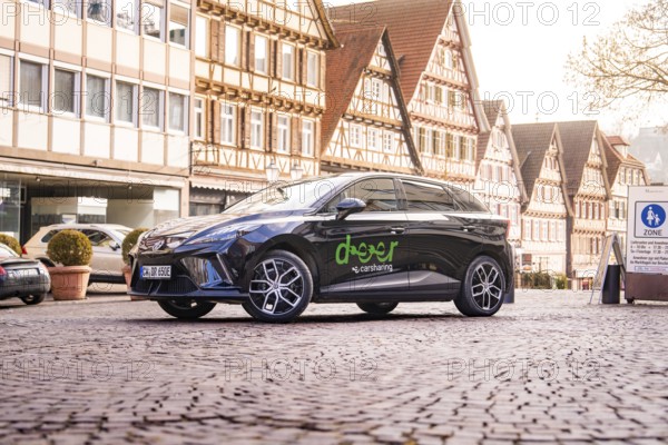 A black car on a cobblestone square surrounded by half-timbered houses in a historic old town, MG4 electric car, Deer e-Carsharing, Calw, Germany