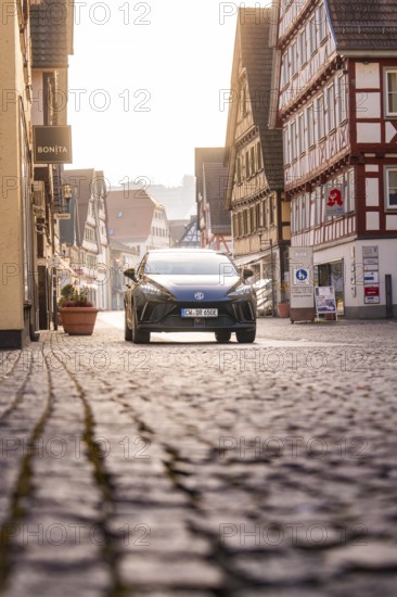 Cobblestone street with a car surrounded by half-timbered houses in glowing sunlight, MG4 electric car, Deer e-Carsharing, Calw, Germany